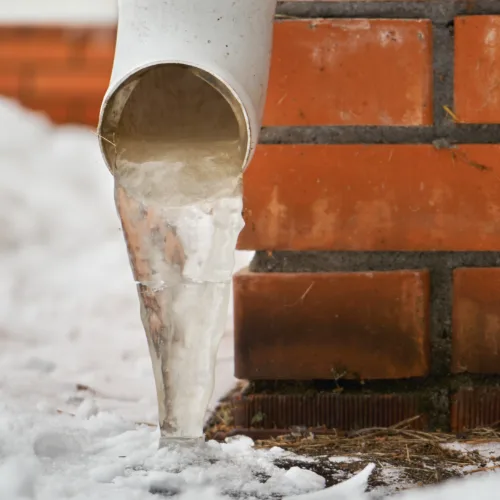A drainpipe on a brick wall with ice coming out of it and snow in the background.